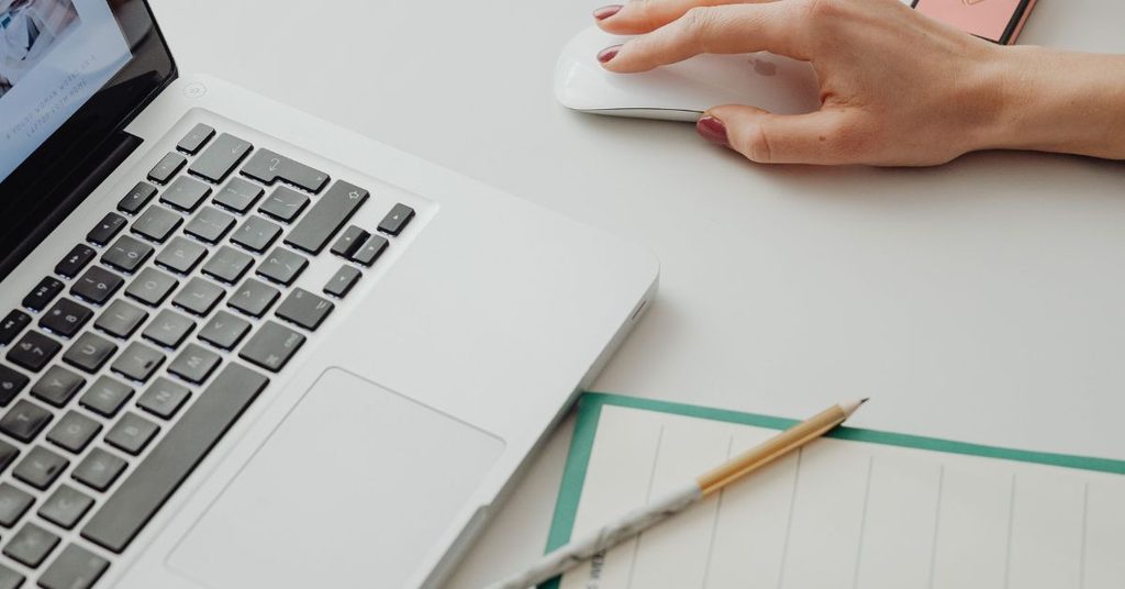 A hand on a mouse beside a laptop and a blank weekly planner pad with a pencil