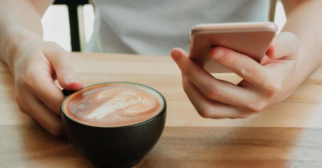 Hands holding a phone and a latte at a wooden table, checking an AI answer before acting on it