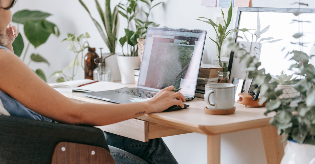Mom typing on a laptop at a wood desk with plants and a coffee mug, using AI to organize her life